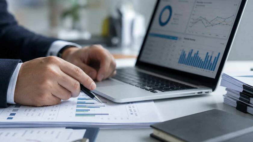 Un professionnel de la finance analyse des graphiques de performance sur un ordinateur portable et des documents imprimés pour structurer une approche par les risques en LCB-FT.