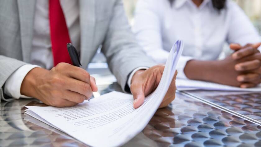 Un homme en costume gris et cravate rouge signe des documents officiels pour valider la conformité réglementaire, assis à une table en métal avec une collègue.