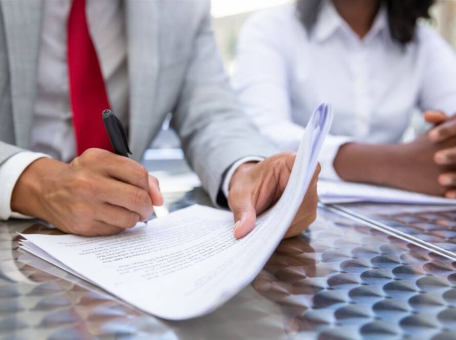 A man in a gray suit and red tie is signing official documents to certify regulatory compliance while sitting at a metal table with a colleague.
