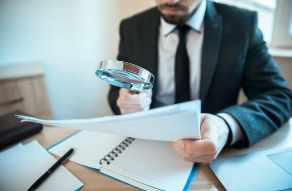 Un homme en costume assis à son bureau examine attentivement des feuilles volantes à la loupe, illustrant l'obligation de documenter et tracer l’EDD lors d'un audit de conformité.