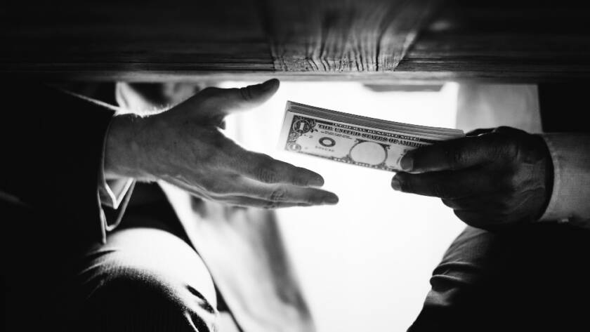 Photo en noir et blanc montrant un échange discret de liasse de billets sous une table en bois, illustrant les transactions illicites liées au financement du terrorisme et blanchiment.