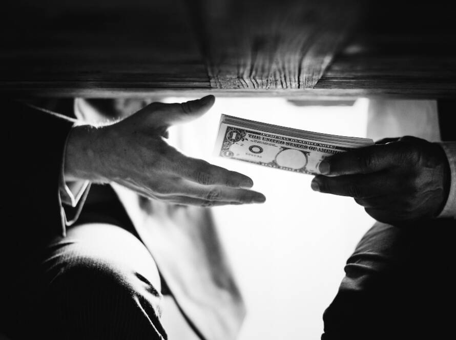 A black-and-white photo showing a discreet exchange of wads of cash under a wooden table, illustrating illicit transactions related to terrorist financing and money laundering.