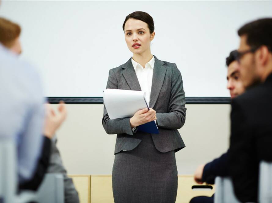 A professional woman in a gray suit holds a folder and speaks to an audience of businessmen at a conference, illustrating the role of a politically exposed person.