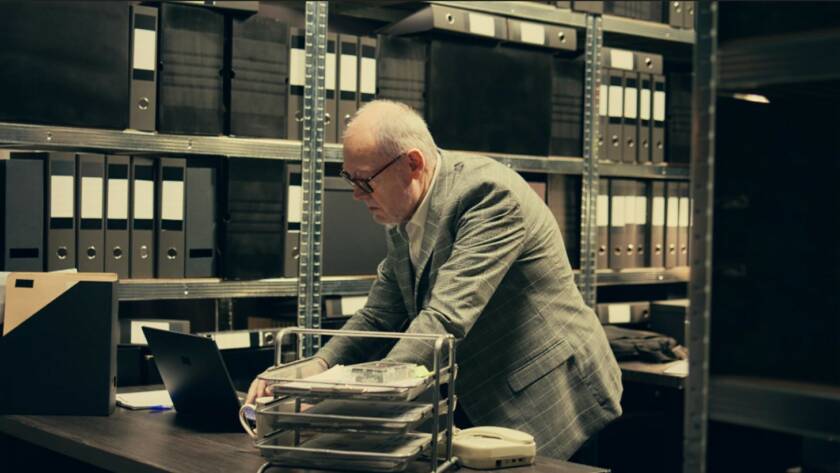 Un expert en conformité âgé, portant des lunettes et une veste de costume grise, examine des documents sur son ordinateur portable dans une salle d'archives remplie de classeurs noirs, illustrant l'application rigoureuse du règlement européen anti-blanchiment.