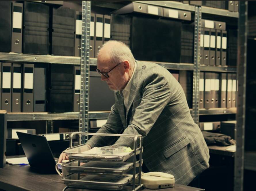 An elderly compliance expert, wearing glasses and a gray suit jacket, reviews documents on his laptop in an archive room filled with black filing cabinets, illustrating the rigorous enforcement of European anti-money laundering regulations.